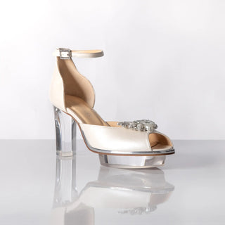 Ein zauberhafter Brautschuh als Opentoe Riemchenpump von der Firma Cinderella’s by Marcela in Ivory/Perlfarbe mit elegantem Nuss-Logo (als Inspiration aus dem Märchen drei Nüsse für Aschenbrödel) sowie hochwertigem transparenten Absatz und Plateau. Die Schuhe sind mit silbernen Accessoires verziert, und die Schnalle ist mit funkelnden Steinchen veredelt. Perfekt geeignet für Hochzeiten sowie alle gehobene Events.