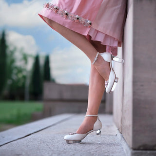 Eine Frau posiert in einem rosa Dirndl (Kleid) von Astrid Söll mit Brautschuhen von Cinderellas_by_Marcela in ivory-Farbe und rosafarbenen Brosche draußen bei einem alten Gebäude.