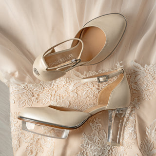 Ein zauberhafter bequemer Brautschuh als Closetoe Riemchenpump von der Firma Cinderella’s by Marcela in Ivory/Perlfarbe mit elegantem Nuss-Logo (als Inspiration aus dem Märchen drei Nüsse für Aschenbrödel) sowie hochwertigem transparenten Absatz und Plateau. Die Schuhe sind mit silbernen Accessoires verziert, und die Schnalle ist mit funkelnden Steinchen veredelt. Perfekt geeignet für Hochzeiten sowie alle gehobene Events.