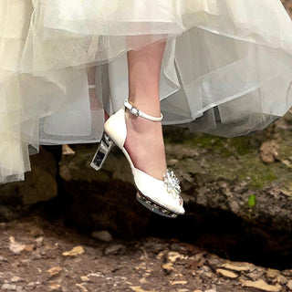 Eine wunderschöne Braut posiert in einem glitzerndem Brautkleid und silbernen CINDERELLAS Brautschuhen aus strukturiertem silber-schimmerndem Leder mit hochwertigem transparenten Absatz und Plateau von Cinderellas by Marcela in einem Park. Die ganze Szenerie sieht wie in einem Märchen aus. Die Braut trägt eine weiße Schleife in den Haaren und schaut runter zu ihrem Cinderella Schuh. Es erinnert an das Märchen Drei Nüsse für Aschenbrödel.