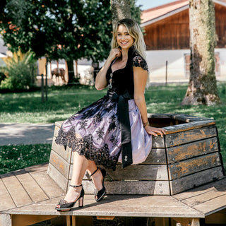 Eine schöne Frau posiert in einem schwarz-lila Dirndl von Astrid Söll mit märchenhaften Cinderellas by Marcela Dirndlschuhen aus schwarzem Wildleder mit einer Blume als Schuhaccessoires auf einer Bank. Es handelt sich um einen exklusiven bayerischen Outfit geeignet für Oktoberfest, Weisswurstparty und alle gehobene Trachtenevents.