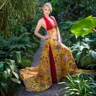 Eine schöne blonde Frau posiert in einem langen rot-gelben Kleid mit märchenhaften Cinderellas Schuhen aus rotem Wildleder mit hochwertigem transparenten Absatz und Plateau mit einem hochwertigen Nuss-Logo von Cinderellas by Marcela in einem botanischen Garten. Ihre Haare sind nach hinten gebunden und sie trägt in den Haaren eine rote Blume.