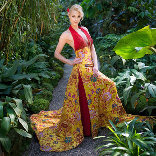 Eine schöne blonde Frau posiert in einem langen rot-gelben Kleid mit märchenhaften Cinderellas Schuhen aus rotem Wildleder mit hochwertigem transparenten Absatz und Plateau mit einem hochwertigen Nuss-Logo von Cinderellas by Marcela in einem botanischen Garten. Ihre Haare sind nach hinten gebunden und sie trägt in den Haaren eine rote Blume.