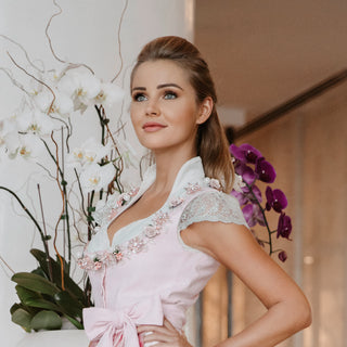 Eine schöne blonde Frau posiert in einem rosafarbenen Dirndl von Astrid Söll mit Brautschuhen aus strukturiertem silber-schimmerndem Leder mit hochwertigem transparenten Absatz und Plateau mit einer Hirschgeweihstickerei von Cinderellas by Marcela in einem Hotel in Dubai. Es kann sich um einen bayerischen Braut-Outfit handeln.