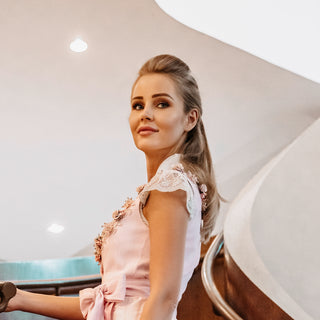 Eine schöne blonde Frau posiert in einem rosafarbenen Dirndl von Astrid Söll mit Brautschuhen aus strukturiertem silber-schimmerndem Leder mit hochwertigem transparenten Absatz und Plateau mit einer Hirschgeweihstickerei von Cinderellas by Marcela in einem Hotel in Dubai. Es kann sich um einen bayerischen Braut-Outfit handeln.