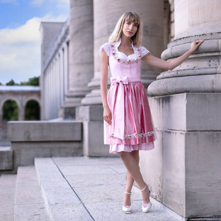 Eine schöne Frau posiert in einem rosa Dirndl (Kleid) von Astrid Söll mit Brautschuhen von Cinderellas_by_Marcela in ivory-Farbe und rosafarbenen Brosche draußen bei einem alten Gebäude.