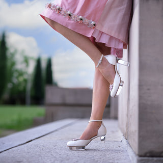 Eine Frau posiert in einem rosa Dirndl (Kleid) von Astrid Söll mit Brautschuhen von Cinderellas_by_Marcela in ivory-Farbe und rosafarbenen Brosche draußen bei einem alten Gebäude.