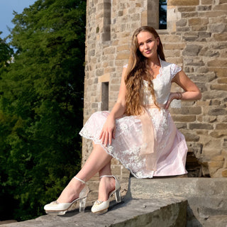 Eine junge Frau sitzt lächelnd auf einer Steinmauer vor einem historischen Gebäude. Sie trägt ein Dirndl von Astrid Söll und elegante Cinderella's Brautschuhe von Bavarian Cinderella mit transparentem Absatz. Im Hintergrund sind grüne Bäume und die steinerne Fassade eines Turms zu sehen, was der Szene eine friedliche und romantische Atmosphäre verleiht.